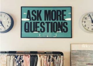 Wall with a poster between two clocks and above a clothes rack. The poster sas "Ask More Questions"