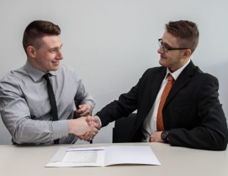 Two young businessmen smiling as they shake hands with a folder of documents open on the table in front of them.